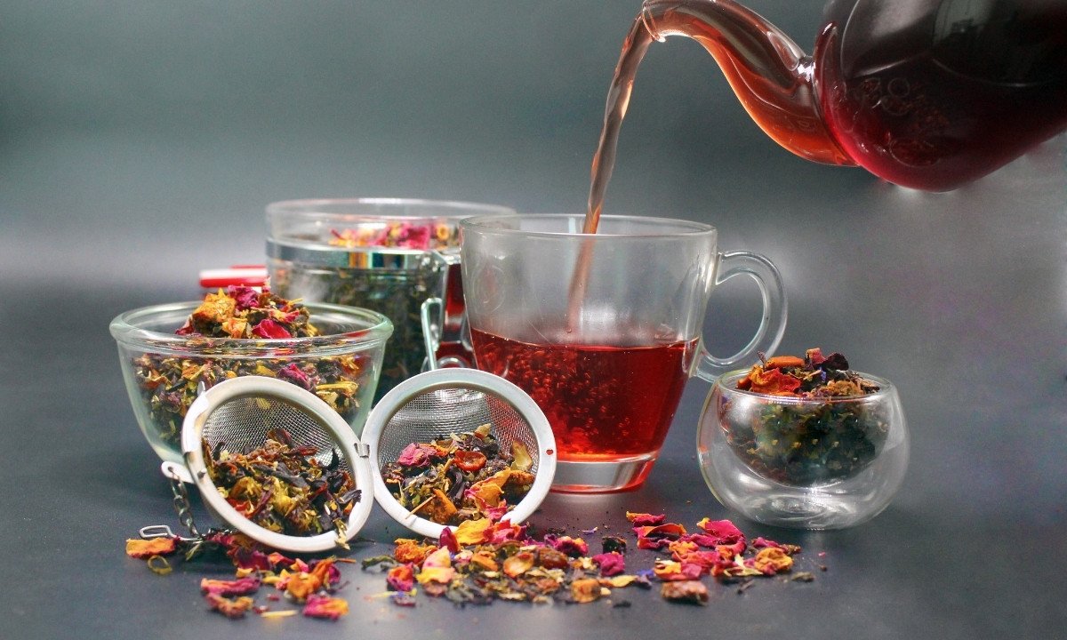 A scene of loose-leaf tea. A glass teapot is pouring hot red tea into a teacup. Next to it are two bowls filled with dried tea leaves, a small glass cup of tea, and an open metal tea infuser overflowing with loose tea leaves. This image illustrates the use of loose-leaf tea as an alternative to tea bags that may contain plastic